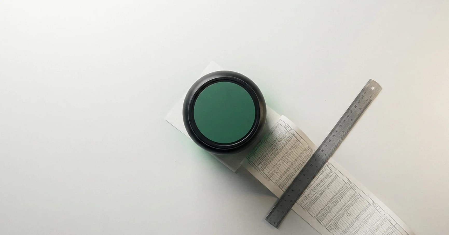 Blick von oben auf eine Dose mit grünem Deckel auf heller Fläche, daneben ein bedruckter Papierstreifen mit Zahlenkolonnen und ein Stahllineal quer darüber.