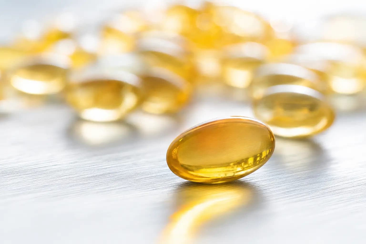 Nahaufnahme einer einzelnen bernsteinfarbenen Softgel-Kapsel auf hellem, gebürstetem Metall; Sonnenlicht fällt durch die Kapsel und wirft einen goldenen Lichtstreifen, dahinter liegen unscharf weitere Kapseln.