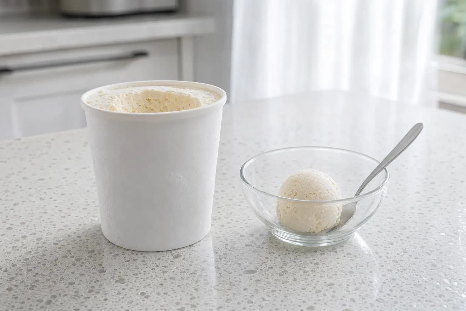 Ein weißer, unbeschrifteter Eisbecher ohne Deckel mit Frost auf einer hellen Terrazzo-Arbeitsplatte, daneben eine kleine Glasschale mit einer einzelnen Kugel Vanilleeis und einem Stahllöffel; im Hintergrund eine helle Küche.